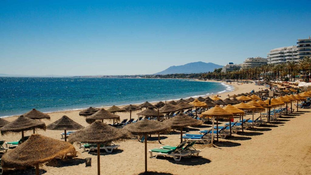 Sunny beach in Marbella, Costa del Sol, with straw parasols, sun loungers, and luxury seafront apartments along the coastline.