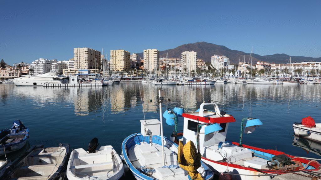 Fishing boats and yachts in a Marbella marina with waterfront buildings and mountains reflected in the calm water.