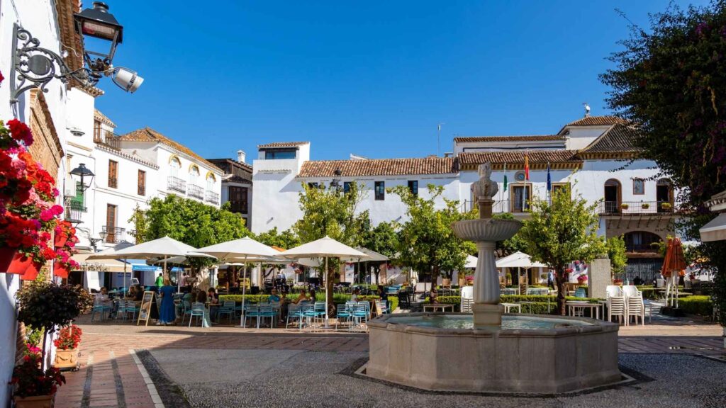 Charming plaza in Marbella Old Town with traditional Andalusian architecture, outdoor cafés, vibrant flowers, and a central fountain under a clear blue sky.