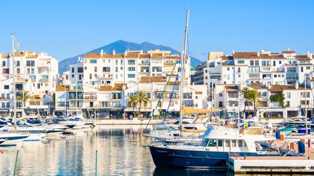 View of luxury yachts and white waterfront buildings in Puerto Banús marina, Marbella, with mountains in the background.