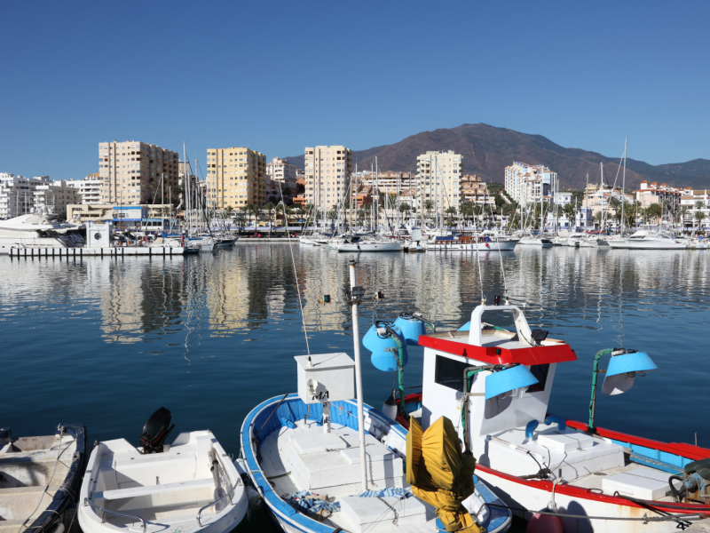 Fishing boats and yachts in a Marbella marina with waterfront buildings and mountains reflected in the calm water.