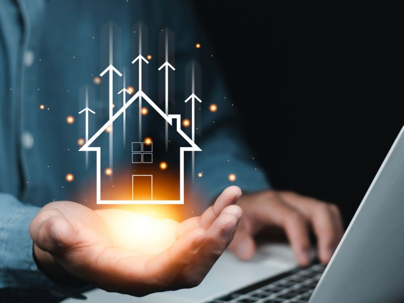 “Person holding a glowing digital house icon with upward arrows, symbolising rising property value and real estate investment growth.”