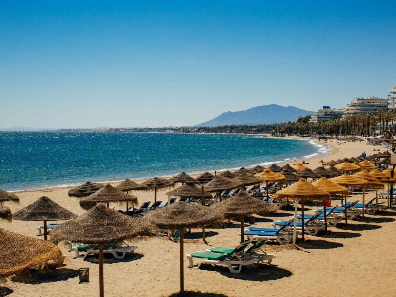 Sunny beach in Marbella, Costa del Sol, with straw parasols, sun loungers, and luxury seafront apartments along the coastline.