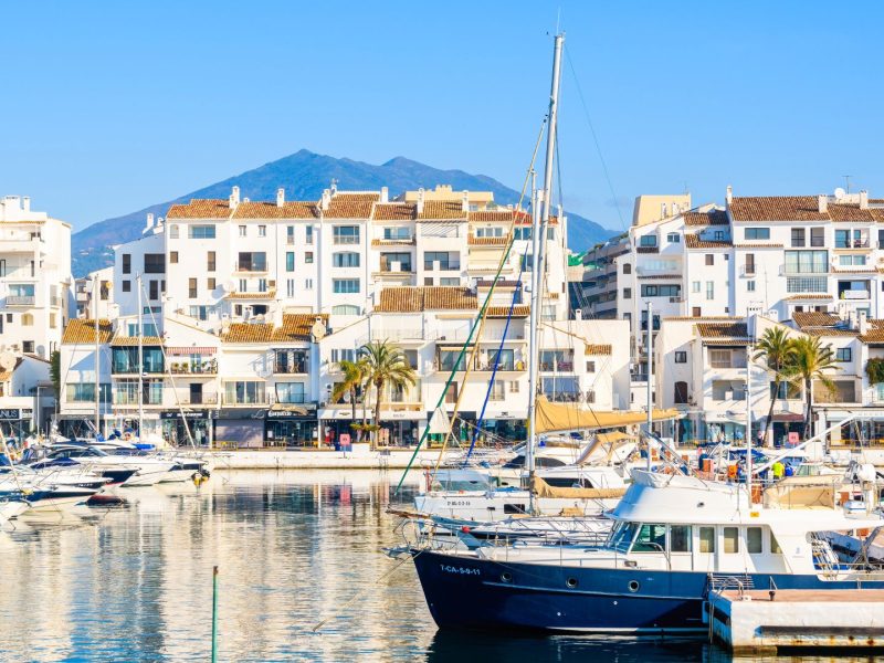View of luxury yachts and white waterfront buildings in Puerto Banús marina, Marbella, with mountains in the background.