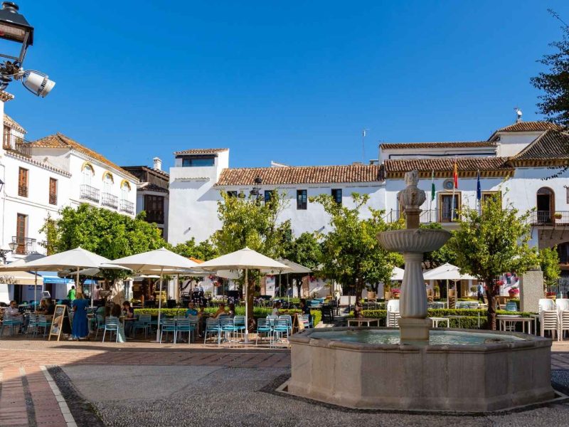Charming plaza in Marbella Old Town with traditional Andalusian architecture, outdoor cafés, vibrant flowers, and a central fountain under a clear blue sky.